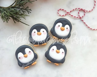 May include: Four black and white penguin cookies with orange beaks and pink cheeks. The cookies are decorated with icing and are arranged on a white surface.