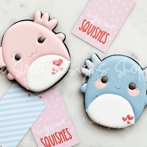 May include: Two decorated sugar cookies shaped like axolotls. The pink axolotl has red hearts on its belly and the blue axolotl has pink hearts on its belly. Both cookies have black eyes and a white belly. The cookies are on a white marble surface with two pink and blue gift tags that say "Hugs and Squishies".