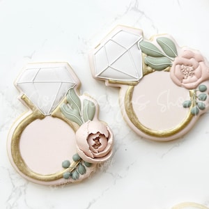 May include: Two sugar cookies decorated with royal icing. One cookie is shaped like a ring with a diamond and a floral design. The other cookie is shaped like a diamond with a floral design.