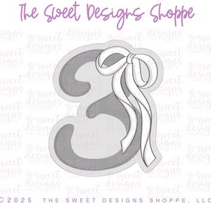 May include: A gray cookie cutter in the shape of the number 3 with a white ribbon bow on top. The cookie cutter is designed for baking and decorating cookies.