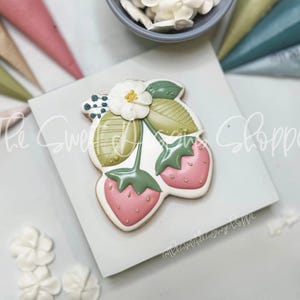 May include: A white cookie decorated with a pink and green strawberry design. The cookie has a white flower with a yellow center and green leaves. The cookie is on a white background.