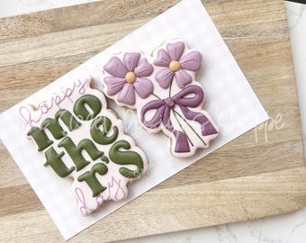 Könnte beinhalten: Zwei dekorierte Zuckerkekse. Ein Keks ist rosa mit grüner Aufschrift "Happy Mother's Day". Der andere Keks ist mit einem Strauß lila Blumen mit einer lila Schleife verziert.