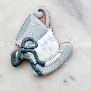 May include: A blue and white teacup and saucer cookie decorated with a blue ribbon. The cookie is decorated with a floral pattern.