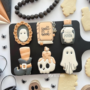 May include: A collection of Halloween-themed cookies on a black tray. The cookies include a ghost in a frame, a spell book, a potion bottle, a witch's boot, a skull candle, and a ghost. The cookies are decorated with orange, white, and gray icing.