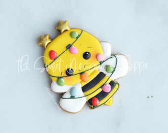 May include: A yellow and black bee cookie decorated with colorful icing and green lines resembling Christmas lights. The bee has a smiling face and large black eyes.