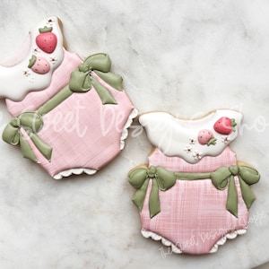 May include: Two baby onesie-shaped cookies. The cookies are pink with a textured pattern and green bows. Each cookie has a white collar and is decorated with strawberries and small white flowers. The cookies are on a white marble surface.