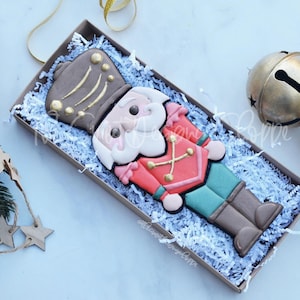 May include: A gingerbread cookie decorated to look like a nutcracker soldier. The cookie is brown, red, green, and white. It is in a box with white shredded paper.