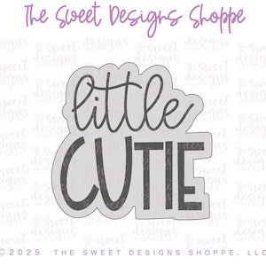 May include: A gray cookie cutter in the shape of the words "little cutie" in a playful font. The text is in a dark gray color, and the cutter has a scalloped outline. The Sweet Designs Shoppe, LLC is printed at the bottom.