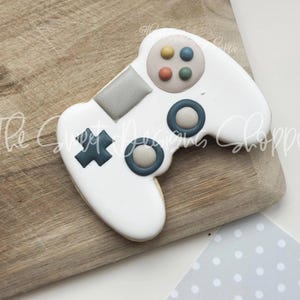May include: White cookie decorated to look like a video game controller. The controller has blue and gray buttons and a black cross.