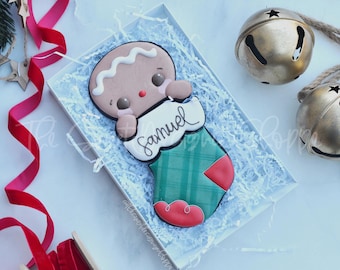 May include: A gingerbread man cookie decorated with white icing and red and brown details, holding a white stocking with the name "Samuel" written in black icing. The cookie is in a white box with white shredded paper.