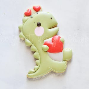 May include: A light green dinosaur-shaped cookie decorated with pink cheeks, a brown eye, and a red heart. Two small red hearts are on top of the dinosaur's head. The cookie is on a white surface.