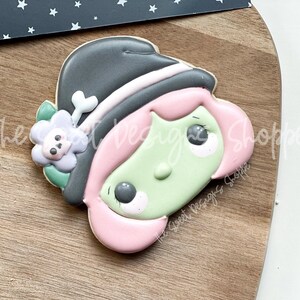 May include: A green-skinned witch cookie with pink hair, a black hat, and a purple flower with a skull bone on the side.
