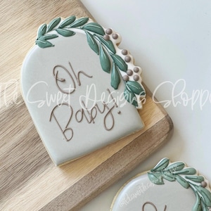 May include: A light gray cookie decorated with green leaves and brown dots. The cookie is shaped like an arch and has the text "Oh Baby!" written in brown cursive.