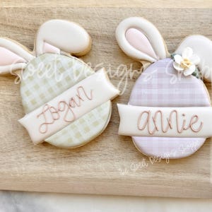 May include: Two decorated Easter bunny cookies with gingham patterns. One cookie is green and says "Logan" on a banner. The other cookie is purple and says "Annie" on a banner.