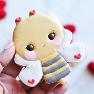 May include: A yellow and gray bee cookie decorated with red hearts. The bee has a cute cartoon face with big eyes and a smile. The cookie is perfect for Valentine's Day.
