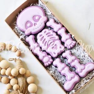 May include: A set of purple skeleton cookies in a box with shredded paper. The cookies are shaped like a skeleton with a head, rib cage, arms, and legs.