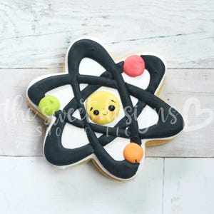May include: A white cookie decorated with black icing in the shape of an atom. The atom has a yellow smiling face in the center and colorful dots around it.