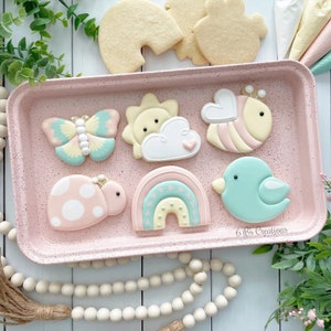 May include: A pink baking sheet with nine decorated sugar cookies. The cookies feature a rainbow, a ladybug, a butterfly, a sun and cloud, a bee, and a blue bird. The cookies are decorated with pastel colors and white icing.
