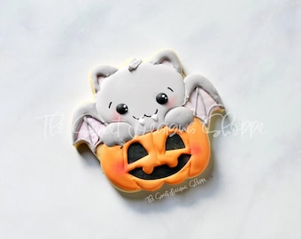 May include: A gray bat cookie with a cute face is perched inside a decorated orange pumpkin cookie. The cookies are decorated with royal icing.