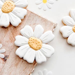May include: White sugar cookies decorated with yellow centers and white icing, resembling daisies. The cookies are on a wooden surface.