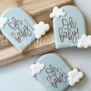 May include: Three blue and white sugar cookies decorated with white clouds and brown icing. The cookies are shaped like arches and have the text "Oh baby!" written on them.