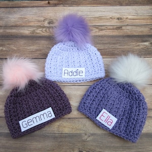May include: Three crocheted hats with pom-poms in purple, pink, and white. Each hat has a personalized name tag: Addie, Gemma, and Ella.