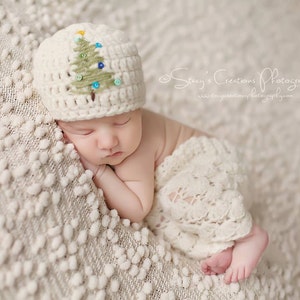 May include: A newborn baby wearing a white crocheted hat with a green and blue embroidered Christmas tree design, sleeping on a white textured blanket.