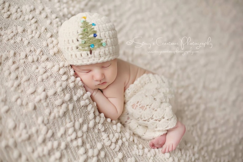 May include: A newborn baby wearing a white crocheted hat with a green and blue embroidered Christmas tree design, sleeping on a white textured blanket.