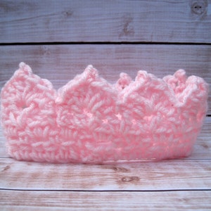 May include: A pink crocheted crown with a pointed top and scalloped edges. The crown is made with a delicate stitch pattern and is perfect for a baby or toddler.