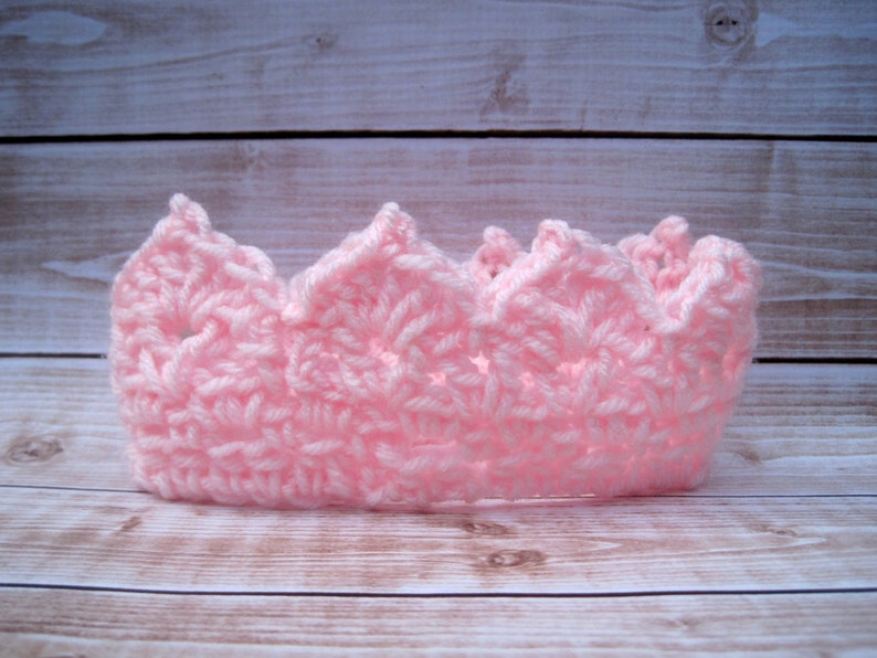 May include: A pink crocheted crown with a pointed top and scalloped edges. The crown is made with a delicate stitch pattern and is perfect for a baby or toddler.
