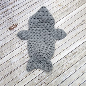 May include: Grey crocheted baby sleeping bag shaped like a fish. The sleeping bag has a hood, fins, and a tail.