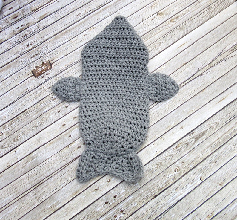 May include: Grey crocheted baby sleeping bag shaped like a fish. The sleeping bag has a hood, fins, and a tail.