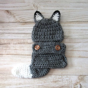 May include: Gray crocheted baby outfit with a wolf hat and a tail. The hat has black ears and the tail is white. The outfit has two brown buttons.