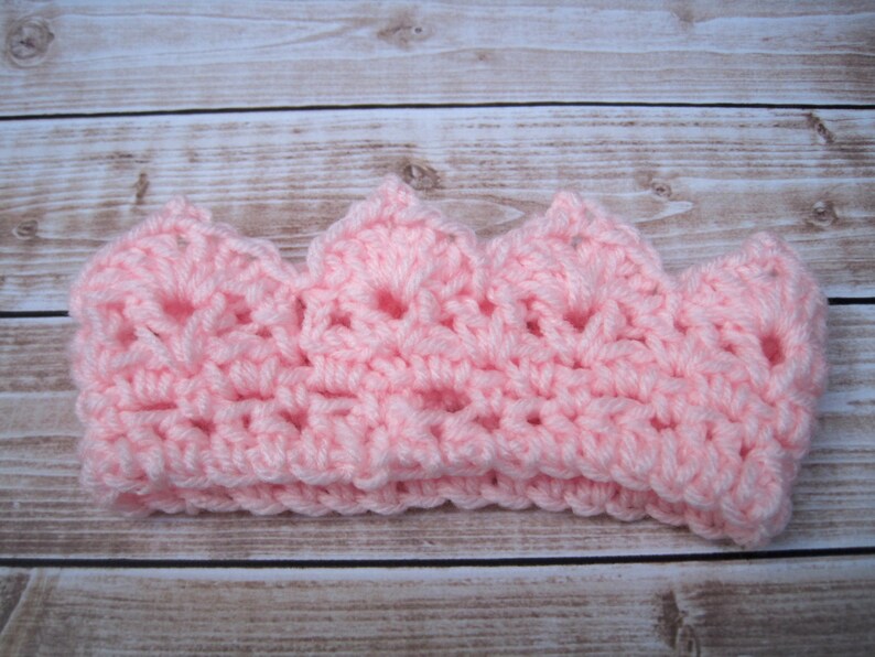 May include: A pink crocheted crown headband. The crown is made with a delicate open stitch pattern.