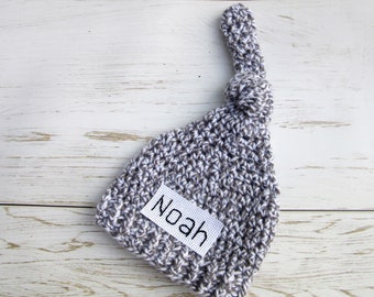 Könnte beinhalten: Eine braune und weiße gehäkelte Baby-Mütze mit einem Knoten oben und einem weißen Etikett mit der Aufschrift "Noah".
