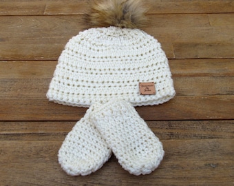 May include: White crocheted hat with a faux fur pom pom and matching mittens. The hat has a brown leather tag that says "Handmade by Jackie".