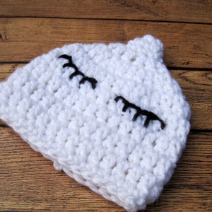May include: A white crocheted baby hat with black embroidered eyelashes. The hat is made of a chunky yarn and has a small, rounded top. The hat is designed to look like a sleeping face.