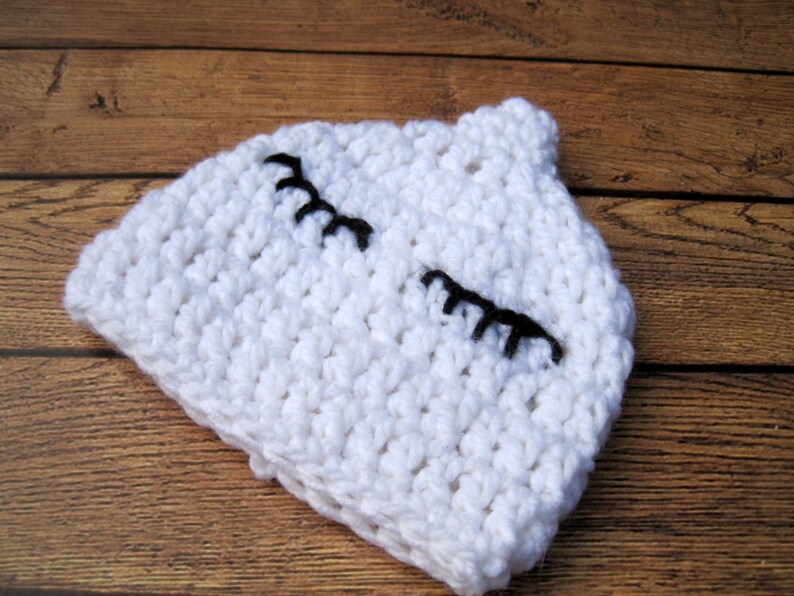 May include: A white crocheted baby hat with black embroidered eyelashes. The hat is made of a chunky yarn and has a small, rounded top. The hat is designed to look like a sleeping face.