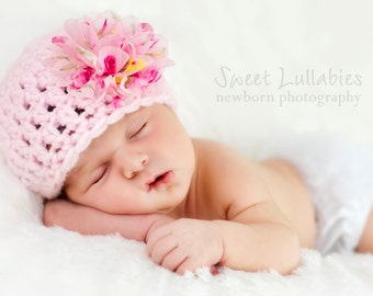 Op de afbeelding: Een slapende baby draagt ​​een roze gebreide muts met een roze bloem erop. De baby ligt op een wit deken. De tekst "Sweet Lullabies newborn photography" staat op de achtergrond.