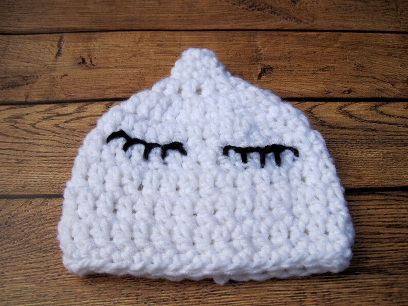 May include: A white, crocheted baby hat with black eyelashes. The hat is handmade and has a textured appearance. The hat is designed to fit a baby's head and is a soft, comfortable accessory.