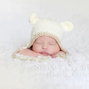 May include: A newborn baby wearing a cream-colored crocheted hat with bear ears, resting on a soft, white, textured surface. The baby's eyes are closed, and the mouth is slightly open. The image is a professional photograph, with the photographer's name at the bottom.
