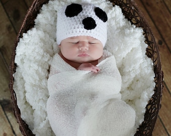 May include: A white crocheted hat with three black circular patches resembling a ghost face, worn by a newborn wrapped in a white swaddle blanket. The infant is resting inside a dark brown woven basket lined with a soft white textured fabric.