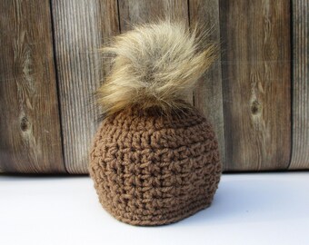 May include: A brown crocheted beanie with a faux fur pom-pom on top. The hat is made of a textured yarn and has a rounded shape. The pom-pom is a light brown color and adds a touch of flair to the hat.
