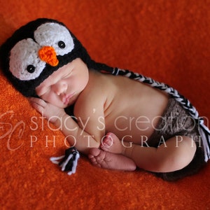 May include: A newborn baby wearing a black and white crocheted penguin hat with an orange beak. The baby is sleeping on a bright orange fuzzy blanket.