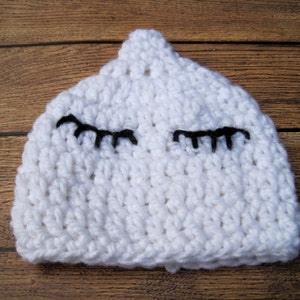 May include: A white crocheted baby hat with black eyelashes. The hat is handmade and has a rounded top. The eyelashes are stitched onto the front of the hat, giving it a sleepy or whimsical appearance.