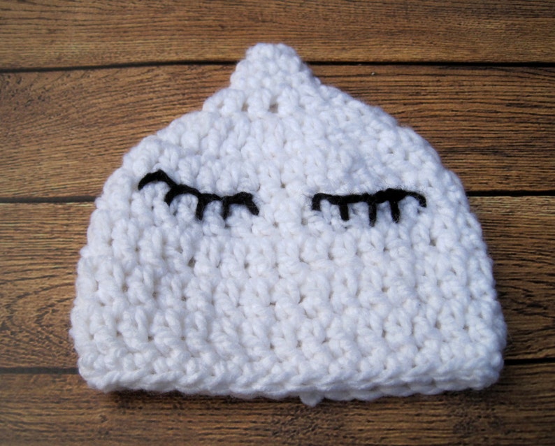May include: A white crocheted baby hat with black eyelashes. The hat is handmade and has a rounded top. The eyelashes are stitched onto the front of the hat, giving it a sleepy or whimsical appearance.