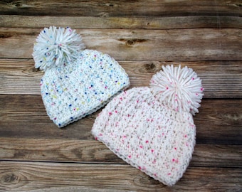 Könnte beinhalten: Zwei weiße Häkelmützen mit bunten Punkten und flauschigen weißen Bommeln. Eine Mütze hat blaue, gelbe und rosa Punkte, während die andere rosa Punkte hat.
