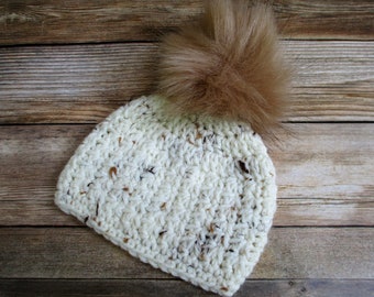 May include: A white crocheted beanie with a brown faux fur pom pom. The beanie has a speckled pattern.