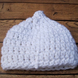 May include: A white, handmade crochet hat. The hat is made of thick, textured yarn and has a rounded crown. The hat is sitting on a wooden surface.