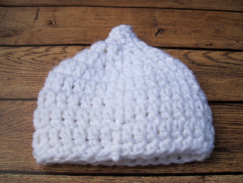 May include: A white, handmade crochet hat. The hat is made of thick, textured yarn and has a rounded crown. The hat is sitting on a wooden surface.